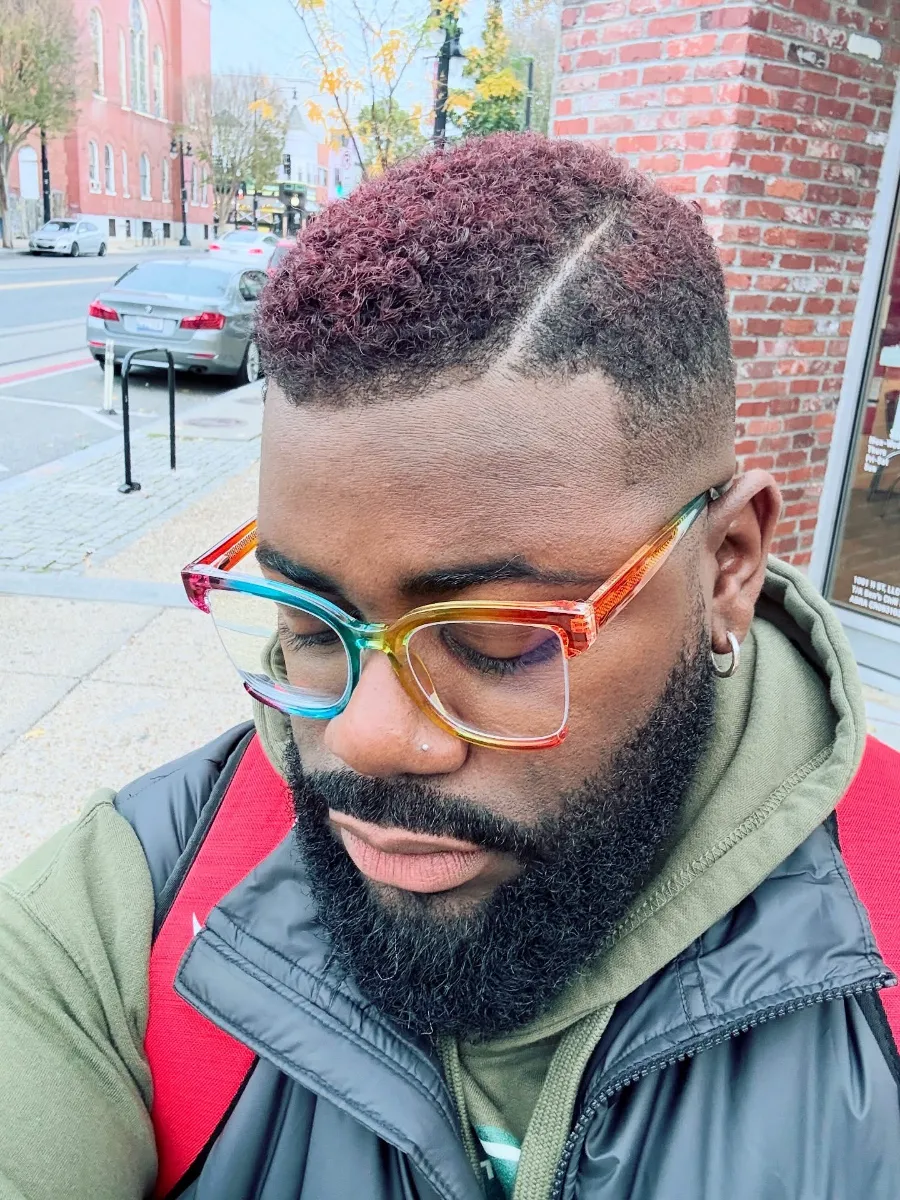 Victor outdoors in rainbow glasses, his short curls colored deep burgundy with a shaved side part and close-faded sides