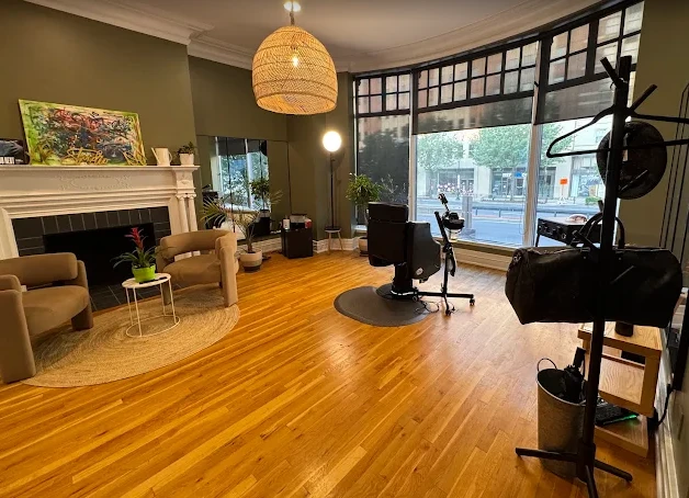 Victor’s studio at XP Studio DC: a sunlit bay window over Connecticut Avenue, one black styling chair on a round mat, wood floors, and a seating corner with two armchairs by a white fireplace